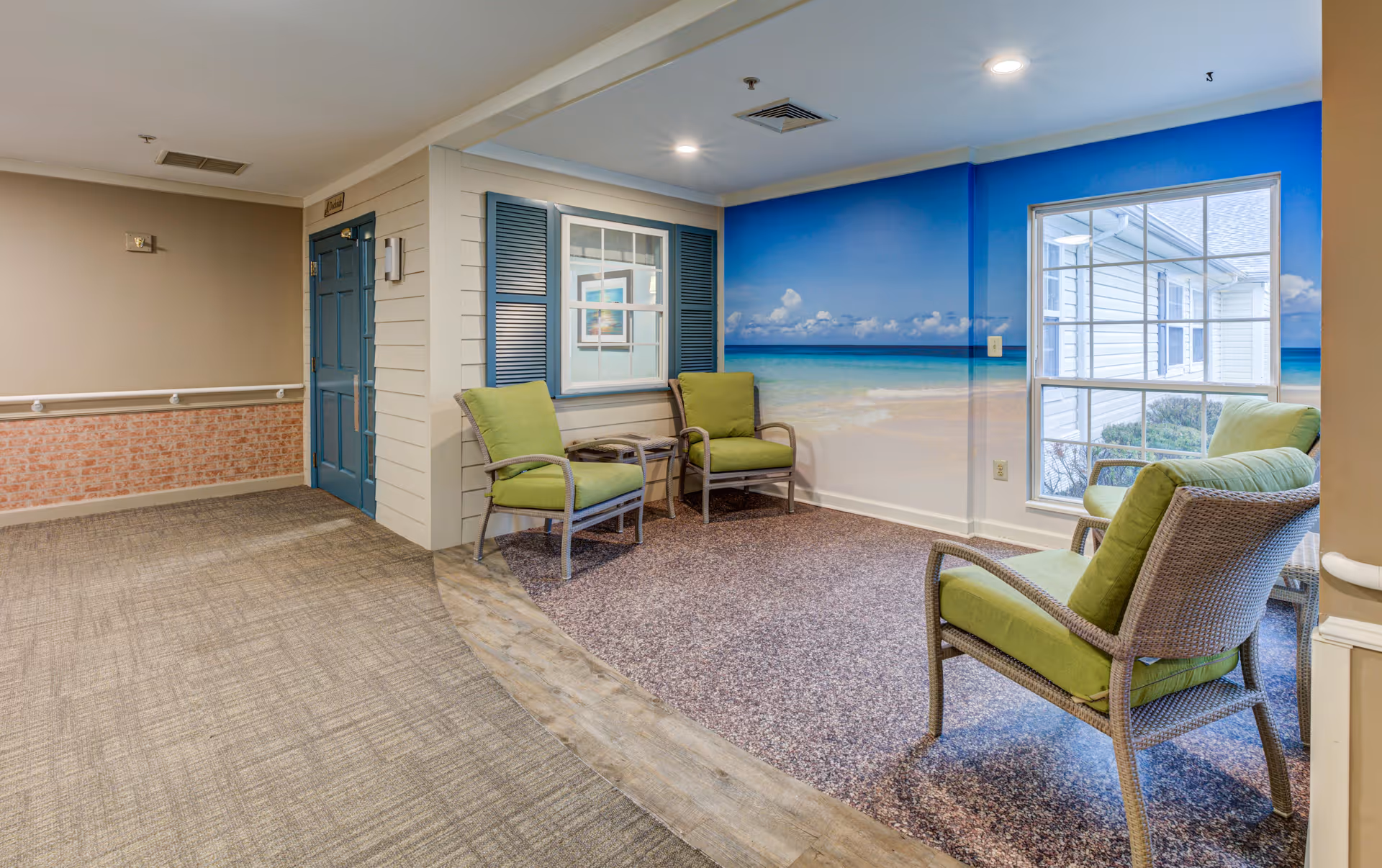 A cozy seating area in a senior living facility hallway featuring four green cushioned chairs arranged around a small table. The walls include a mural of a beach scene with blue skies and ocean waves, and a window with blue shutters. The floor transitions from carpet to a textured surface in the seating nook. The hallway has beige walls with a brick-patterned lower half and a blue door at the end.