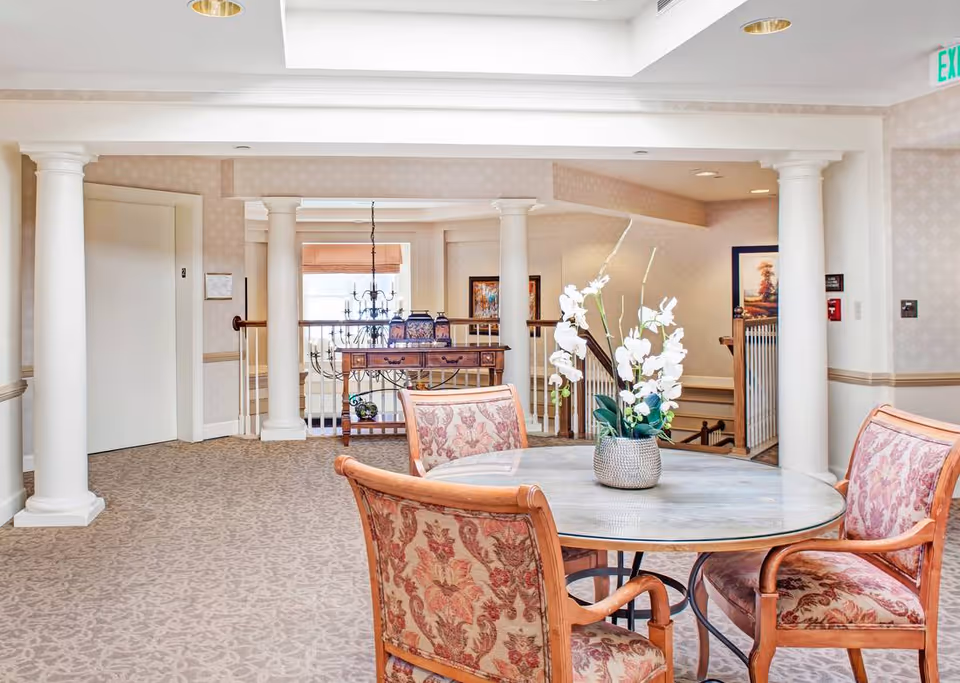 A bright and spacious interior area with a round table and four upholstered chairs featuring floral patterns. A vase with white flowers is placed on the table. The room has beige patterned carpet, white columns, and a console table with decorative items near a railing overlooking a lower level. There is an elevator door on the left and an exit sign on the right.