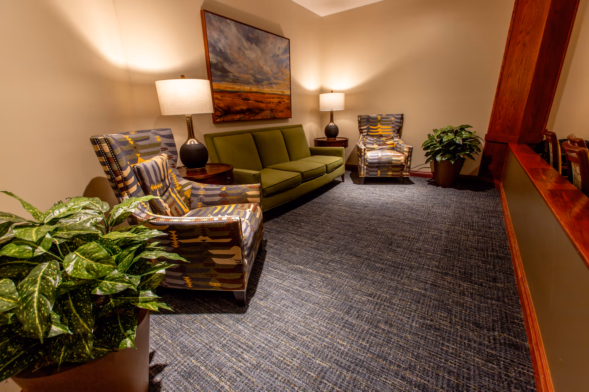 Cozy lounge area with a green sofa, patterned armchairs, table lamps, artwork, and potted plants.