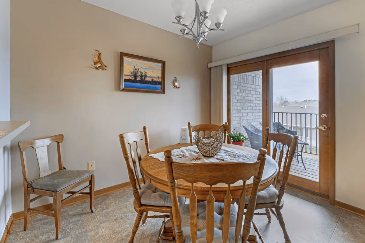 A dining area with a round wooden table surrounded by four wooden chairs with cushions. The table has a decorative centerpiece and a lace table runner. On the wall above the table is a framed painting of a landscape with trees and birds, flanked by two small sailboat wall decorations. To the right, there are glass sliding doors leading to a balcony with two chairs and a small table, overlooking rooftops and trees outside.