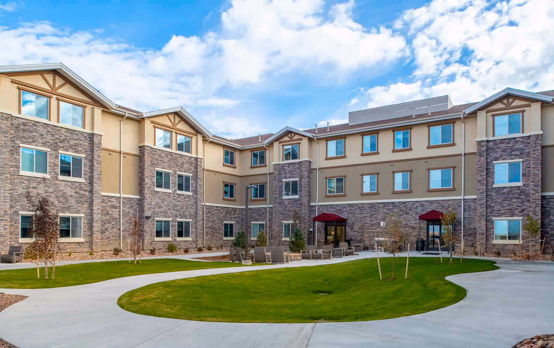 Exterior view of a three-story senior living facility building with stone and beige siding, multiple windows, and two entrances with red awnings. The foreground features a curved concrete walkway surrounding a well-maintained green lawn with small trees and outdoor seating areas.