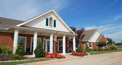 Front entrance of a single-story brick building with white columns, an American flag, and landscaped flower beds under a blue sky.