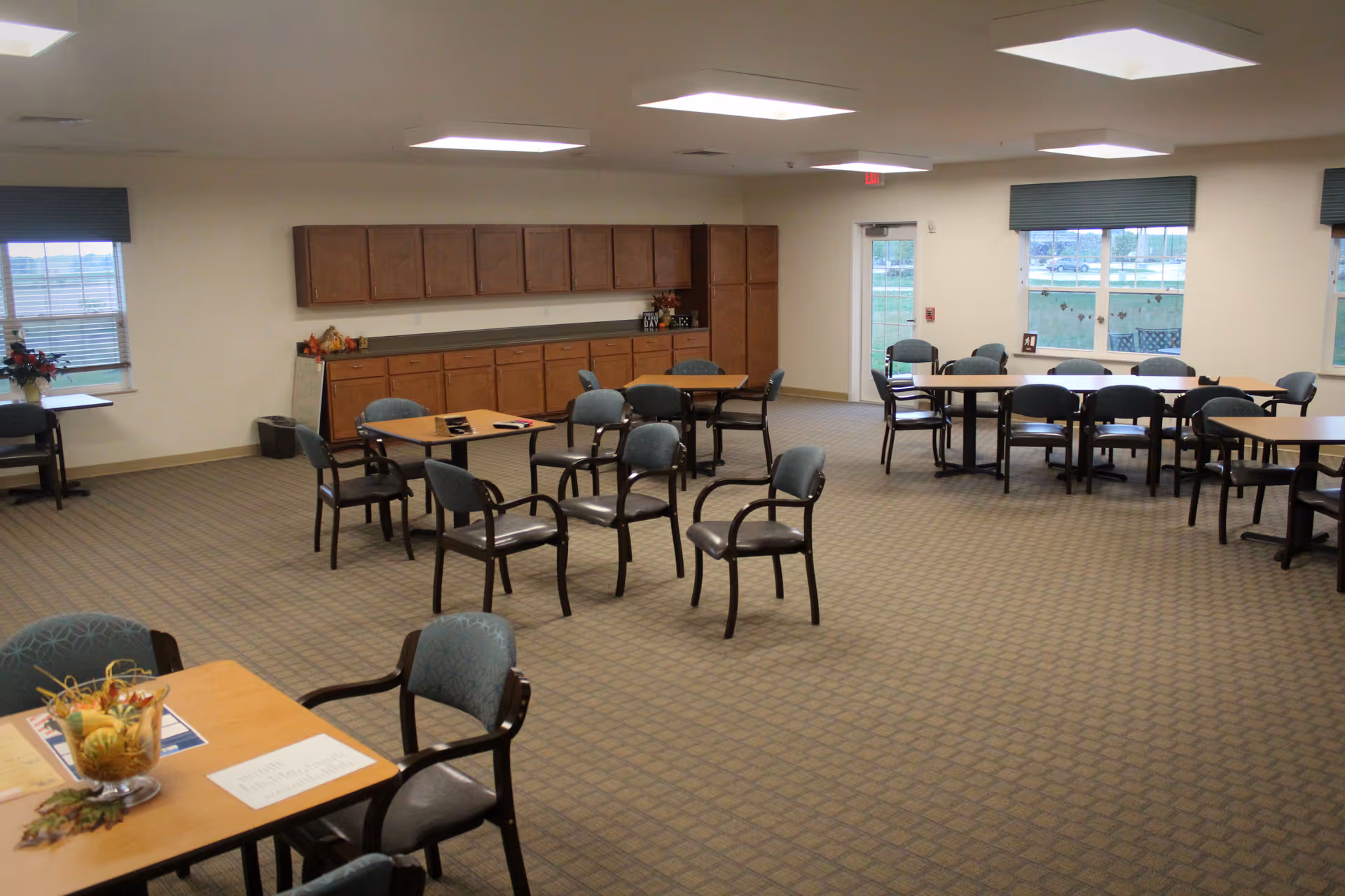 A spacious common area in a senior living facility with multiple tables and chairs arranged for group seating. The room has carpeted flooring, large windows with blinds, overhead fluorescent lighting, and wooden cabinets along one wall. There are some decorative items on the tables and cabinets.