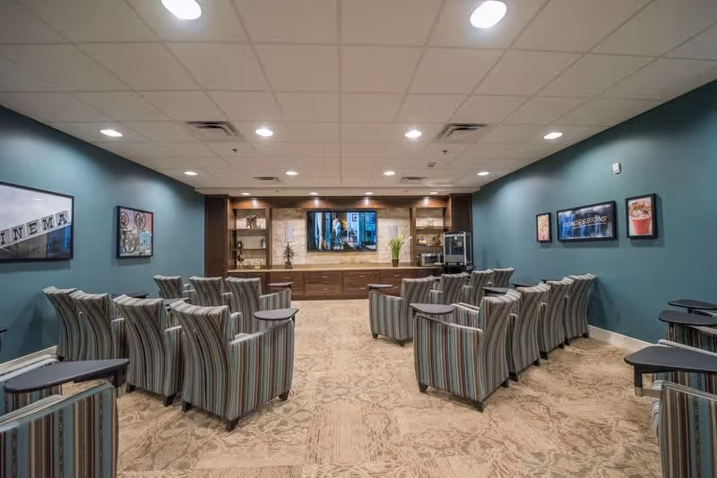 A cozy media room with multiple striped armchairs arranged in rows facing a large flat-screen TV mounted on a wooden entertainment center. The room has teal walls adorned with framed pictures and a carpeted floor with a subtle pattern. The ceiling has recessed lighting.
