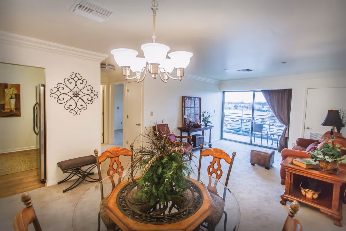 Open-plan apartment interior with a dining table and centerpiece in the foreground, a living area with sofa and side table, and a sliding glass door leading to a balcony.