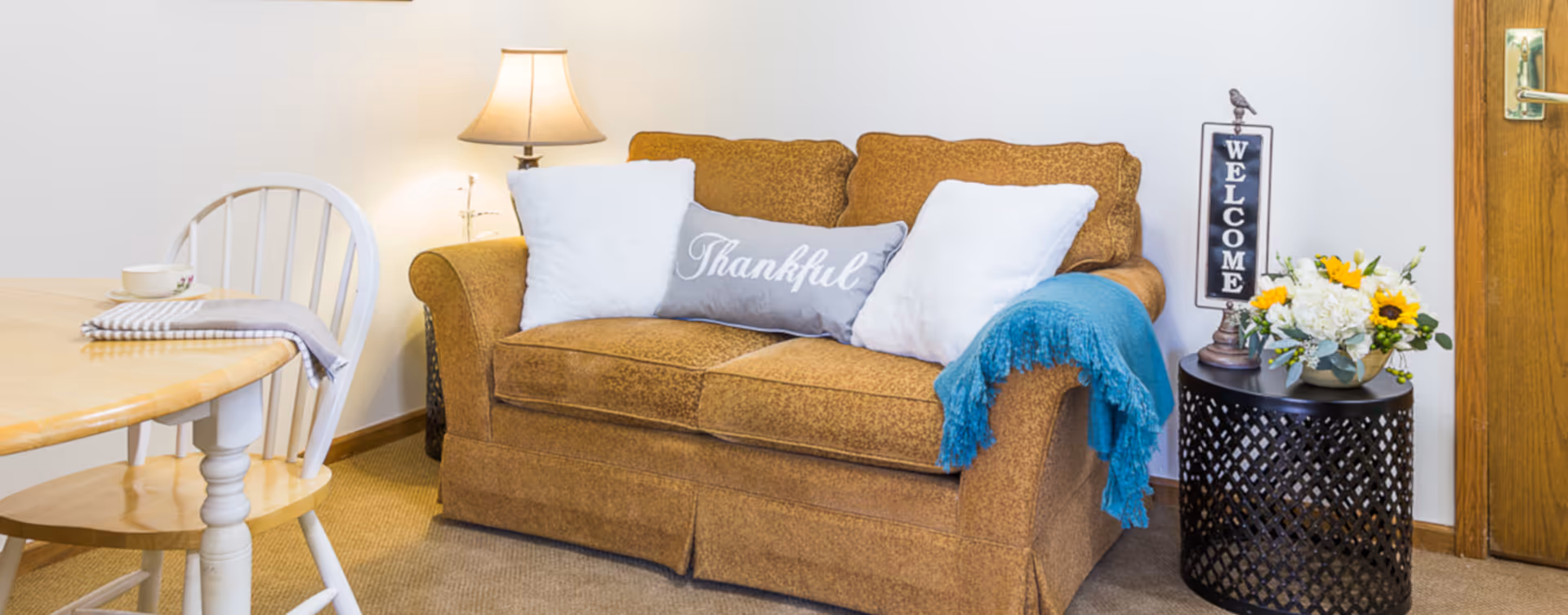 A cozy living room area with a brown upholstered loveseat adorned with two white pillows and a gray pillow with the word 'Thankful'. A blue throw blanket is draped over one arm of the loveseat. To the right, there is a black round side table with a floral arrangement and a decorative sign that says 'WELCOME'. A wooden door with a brass handle is visible in the background. To the left, part of a wooden dining table and a white chair are visible, along with a table lamp providing warm light.