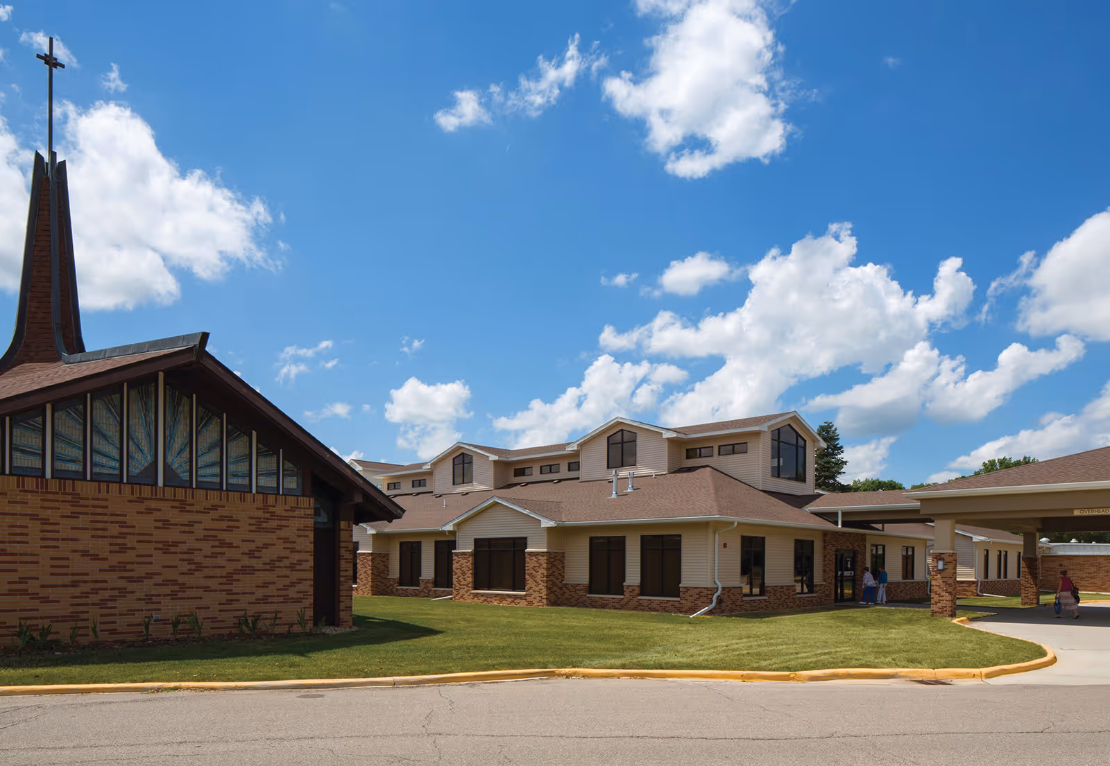 Exterior view of a senior living facility with a brick and beige building under a blue sky with scattered clouds. The building has multiple windows and a covered entrance area. A church-like structure with a tall steeple and cross is visible on the left side.