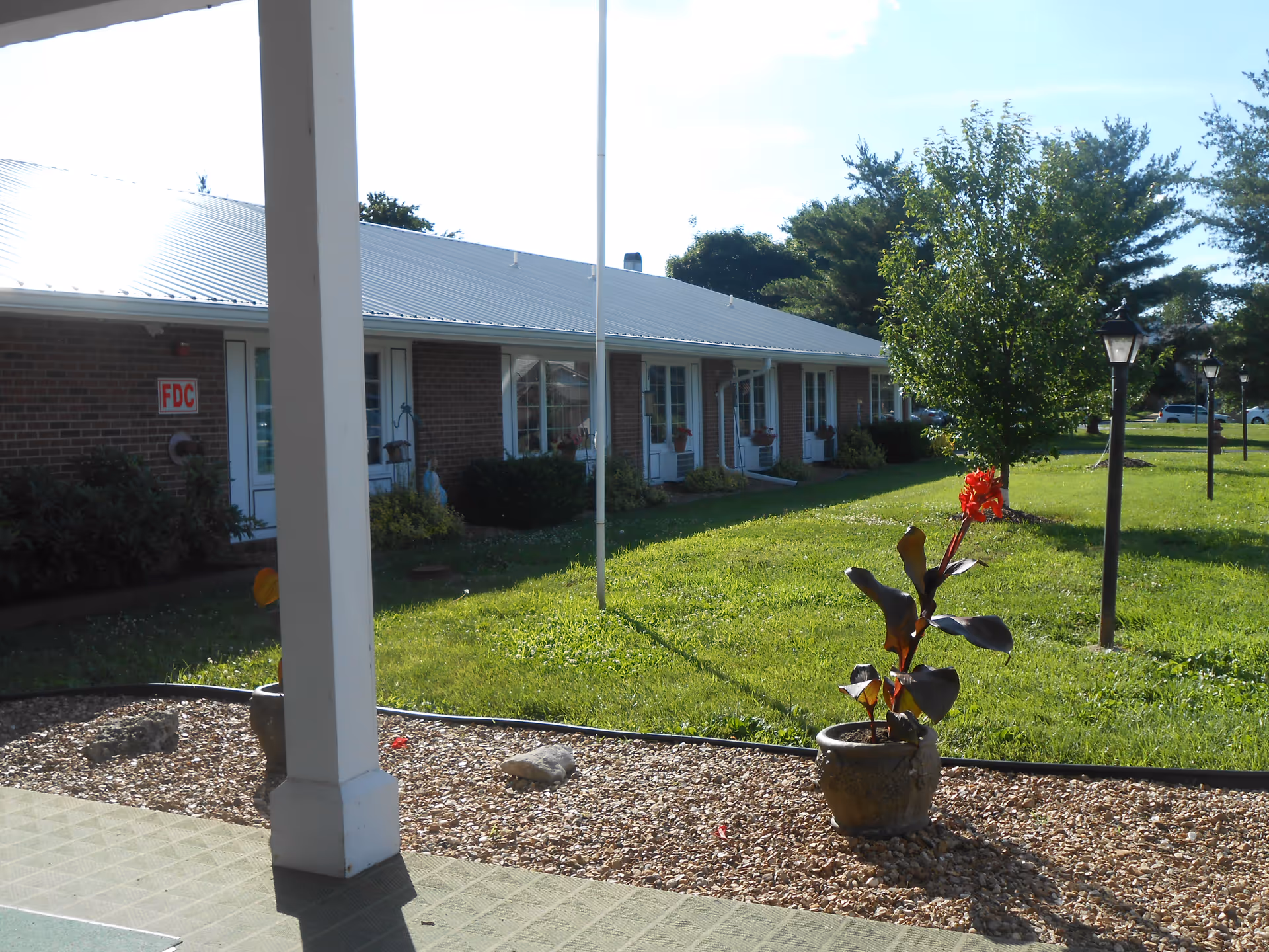 Outdoor view of a single-story brick building with multiple white doors and windows, a metal roof, and a green lawn with trees and lamp posts. A potted plant with red flowers is in the foreground on a gravel bed next to a covered walkway supported by white columns.