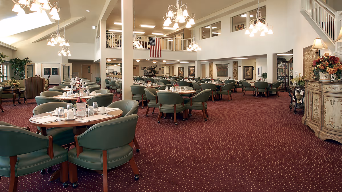 Spacious dining room with multiple round tables set with glasses, napkins, and condiments. The room features green cushioned chairs, red patterned carpet, chandeliers hanging from the ceiling, and an American flag displayed on the upper level balcony. Decorative plants and framed artwork adorn the walls.