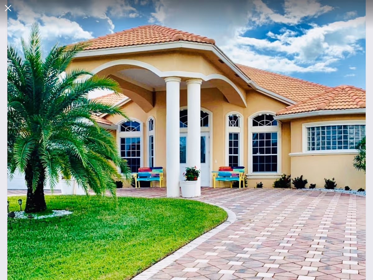 Front exterior view of a single-story building with a tiled roof, large white columns at the entrance, arched windows, a paved driveway, green lawn, and a palm tree under a partly cloudy sky.