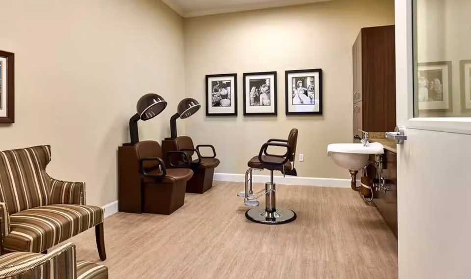 Interior view of a salon area in a senior living facility with two vintage hair dryers, a salon chair, a sink, and two striped armchairs. Three black and white framed photos hang on the beige wall.