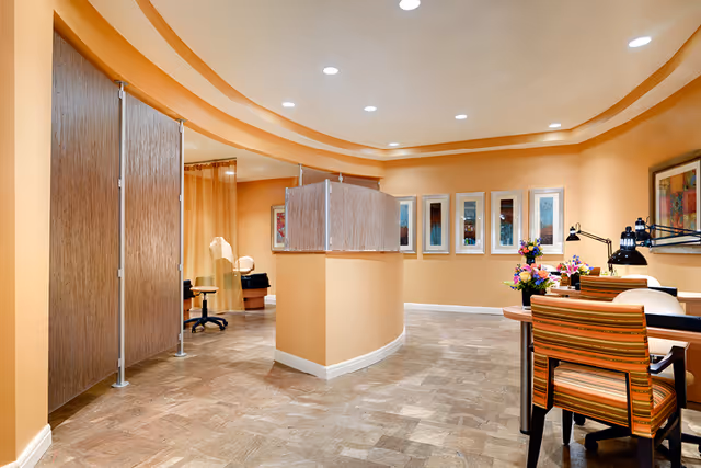 Well-lit peach-toned interior of a senior living salon with manicure stations, chairs, privacy screens, and decorative artwork.