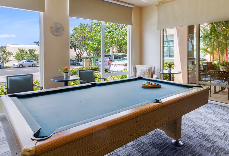 A bright room with a pool table in the center, two small round tables with chairs near large windows showing parked cars and greenery outside. There is also a patio area visible through a glass door with outdoor seating and plants.
