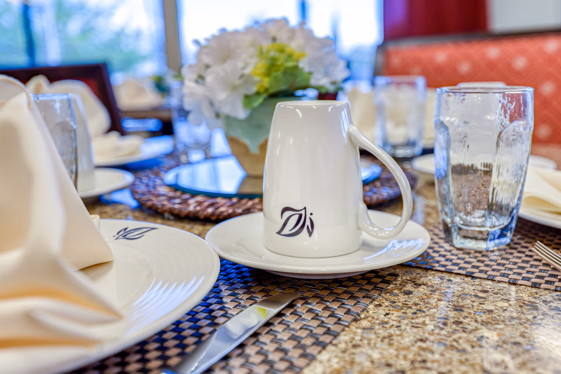 Close-up view of a dining table set with a white coffee cup placed upside down on a saucer, a glass of water, a folded beige napkin on a plate, and silverware. In the background, there is a floral centerpiece and cushioned chairs.