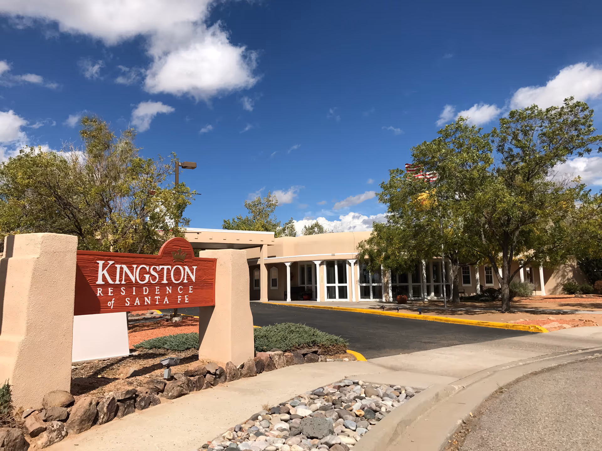 Exterior view of Kingston Residence of Santa Fe, showing the entrance sign in front of a beige building with trees and a clear blue sky with some clouds.