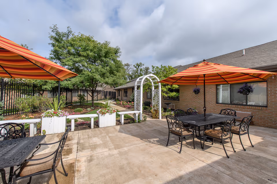 Courtyard patio with metal tables and chairs, striped orange umbrellas, planter boxes and a white arbor beside a brick building.