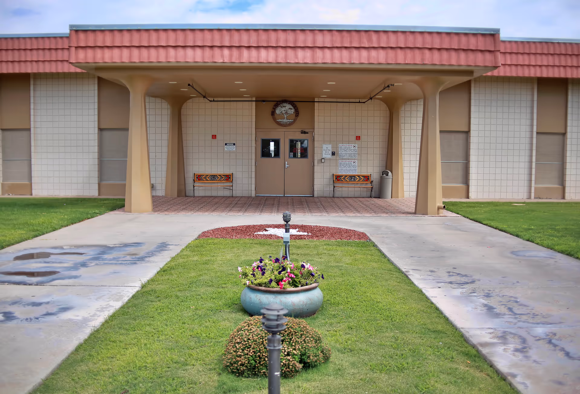Front entrance of Franklin Heights Nursing and Rehab Center showing a covered walkway with two benches on either side of the door, a small garden with flowers and bushes in the foreground, and a building with a tiled facade and a red roof.