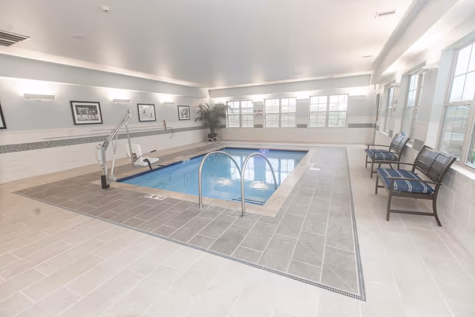 Indoor swimming pool area with tiled floor and walls, a pool lift chair for accessibility, metal handrails, framed pictures on the wall, large windows letting in natural light, and several cushioned benches along the right side.