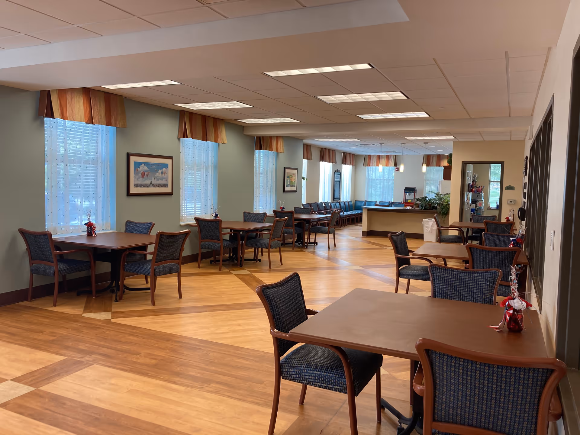 A spacious dining room in a senior living facility with multiple wooden tables and cushioned chairs arranged neatly. The room has large windows with sheer white curtains and valances, allowing natural light to fill the space. The walls are painted light green, and there are framed pictures hanging. In the background, there is a counter area with plants and a popcorn machine.