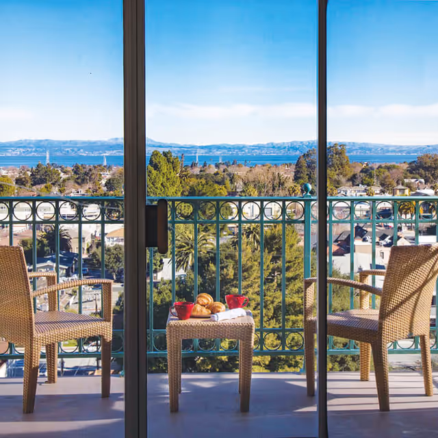 Balcony with two wicker chairs and a small table holding croissants and red cups overlooking treetops and a bay.