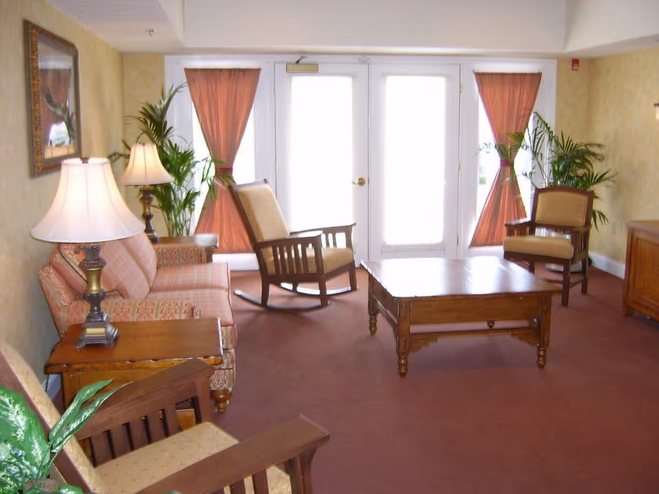 Communal living room with sofas, wooden rocking and arm chairs, a coffee table, lamps, and potted plants in front of glass doors.