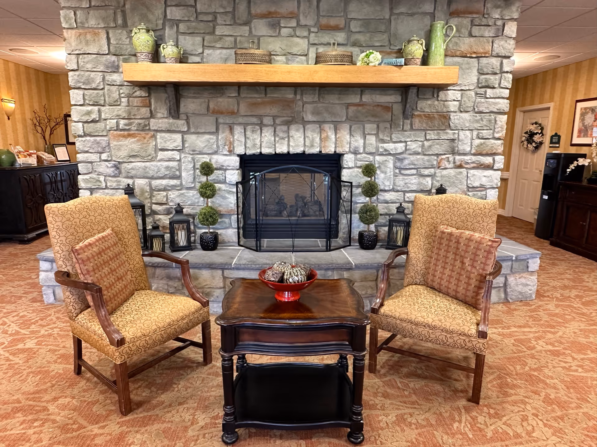 A cozy seating area with two patterned armchairs and a wooden side table in front of a stone fireplace. The fireplace mantel is decorated with green ceramic pitchers and small baskets. The room has warm-toned carpet and walls with subtle striped wallpaper. There are decorative plants and lanterns on the hearth.