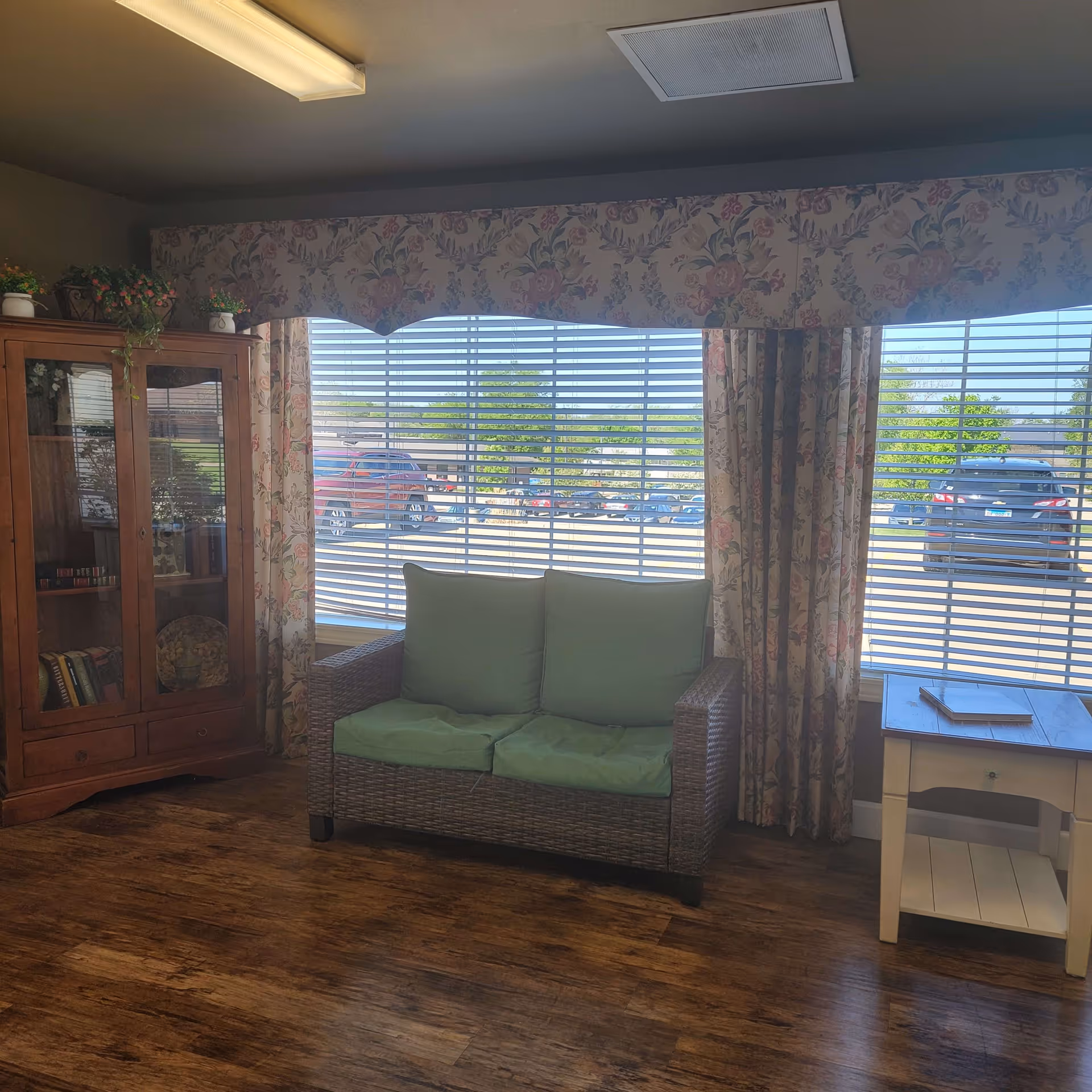 A cozy sitting area with a wicker loveseat featuring green cushions, positioned in front of large windows with horizontal blinds and floral curtains. To the left is a wooden cabinet with glass doors displaying books and decorative items, and to the right is a small white side table with a book on top. The floor is wooden, and the room is softly lit by a ceiling light.