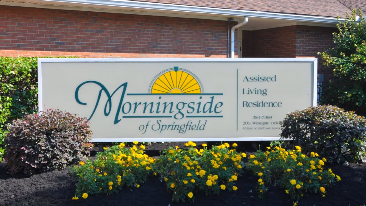 A roadside sign for 'Morningside of Springfield Assisted Living Residence' with a sun logo and yellow flowers in front of a brick building.