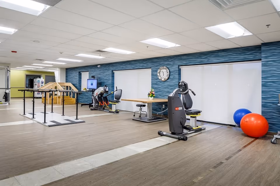 Spacious rehabilitation/exercise room with parallel bars, recumbent exercise bikes, a therapy table, and large stability balls.