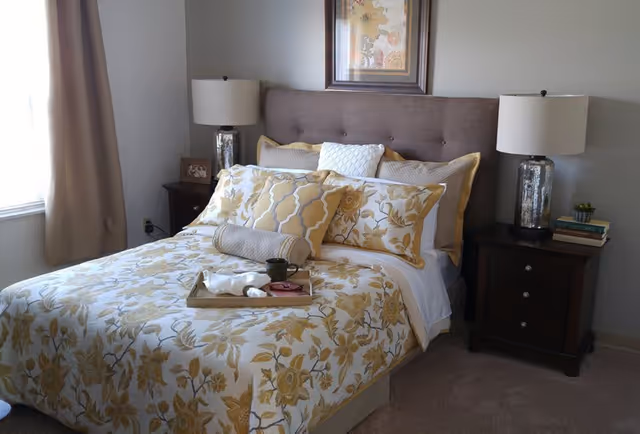 A neatly made bed with a floral patterned bedspread and multiple pillows in shades of yellow and white. The bed has a tufted headboard and is flanked by two dark wooden nightstands, each with a lamp. There is a tray on the bed holding a cup, a napkin, and a small plate. A framed floral artwork hangs above the headboard, and a window with beige curtains is visible on the left side of the room.