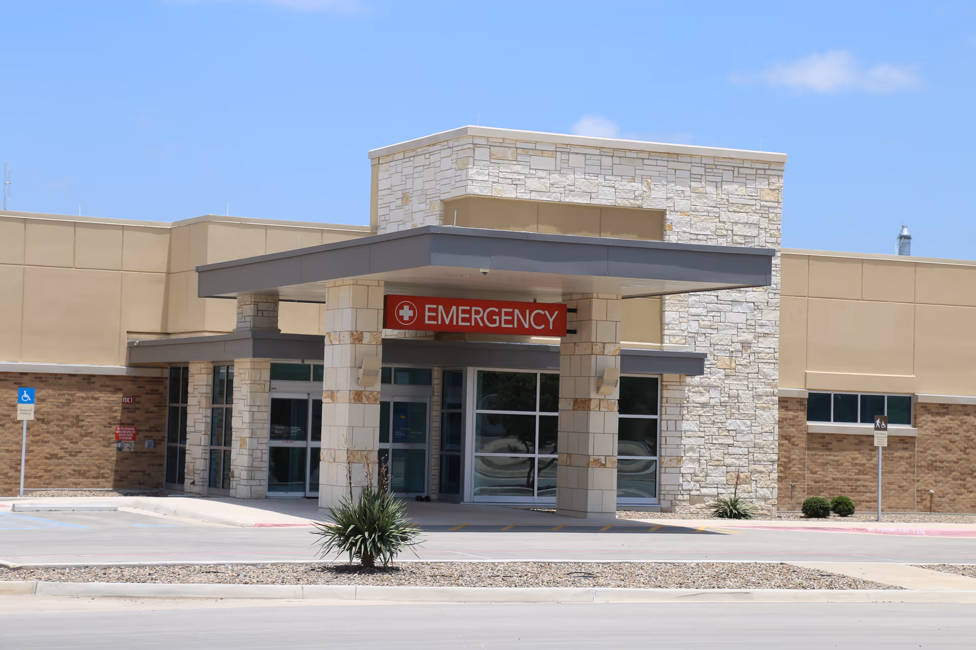 Entrance to the emergency department of a hospital building with a covered drop-off area, stone and beige exterior walls, and a red sign reading 'EMERGENCY' above the doors.