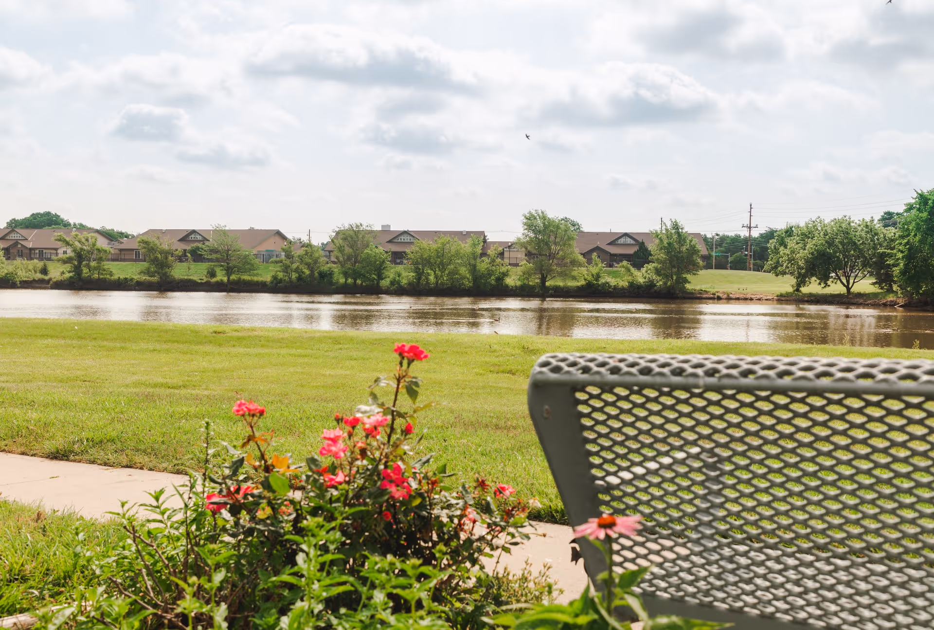 View of a peaceful outdoor area with a metal bench in the foreground, colorful flowers, a grassy lawn, a body of water, and houses with trees in the background under a partly cloudy sky.