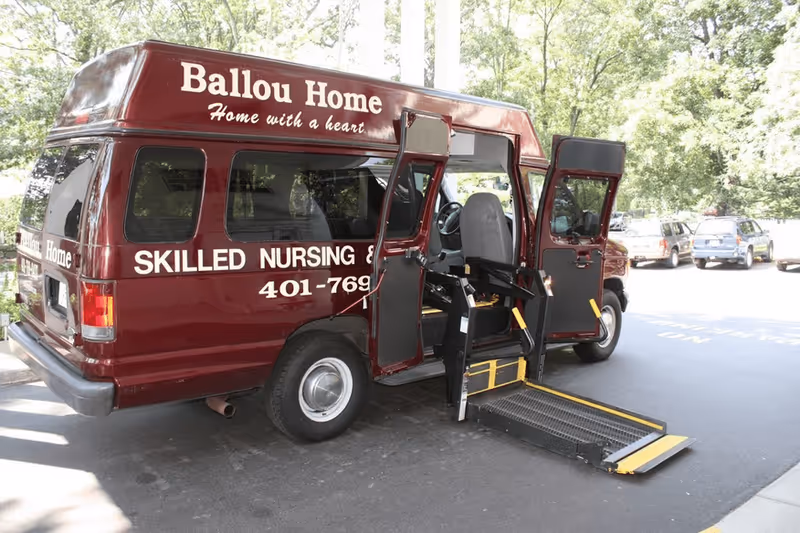 A maroon Ballou Home medical transport van parked outside with its wheelchair lift deployed.