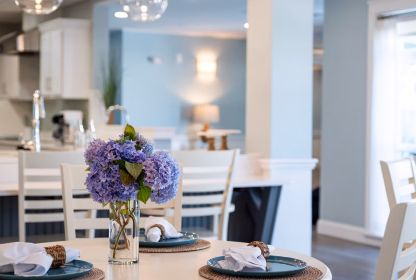 A dining table set with blue plates, white cloth napkins with brown napkin rings, and a clear glass vase holding a bouquet of purple hydrangeas. The background shows a modern kitchen and dining area with white chairs, light blue walls, and soft lighting.