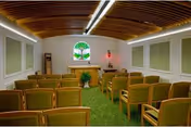Small chapel-style room with rows of wooden chairs facing an altar beneath a stained-glass window and a curved wood-paneled ceiling.