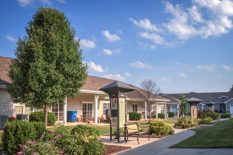 Outdoor view of The Fremont Senior Living facility showing a row of single-story residential buildings with porches, surrounded by well-maintained landscaping including trees, bushes, and flowers under a partly cloudy blue sky. There are benches and informational signposts along a paved walkway.