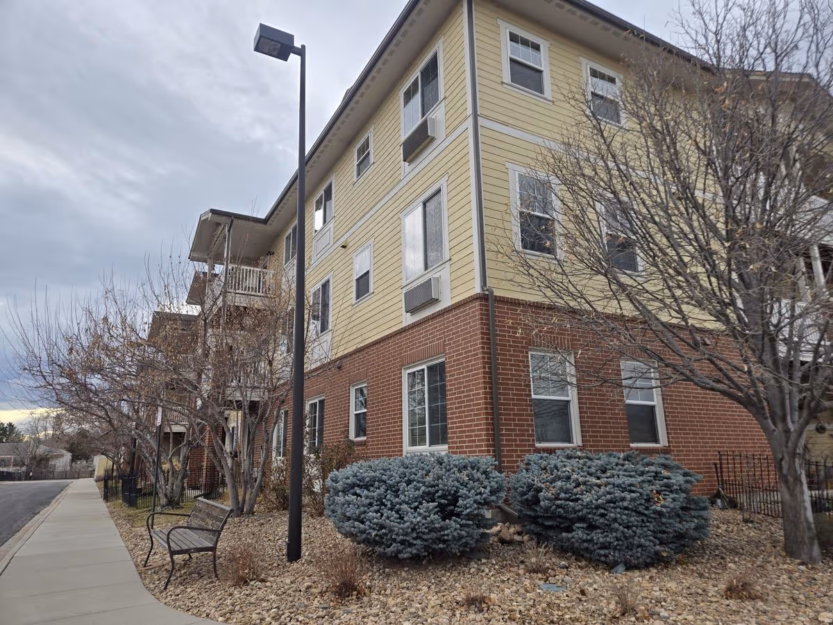 Corner view of a multi-story senior living building with red brick lower levels and yellow siding above, leafless trees, a bench, and a streetlamp along the sidewalk.