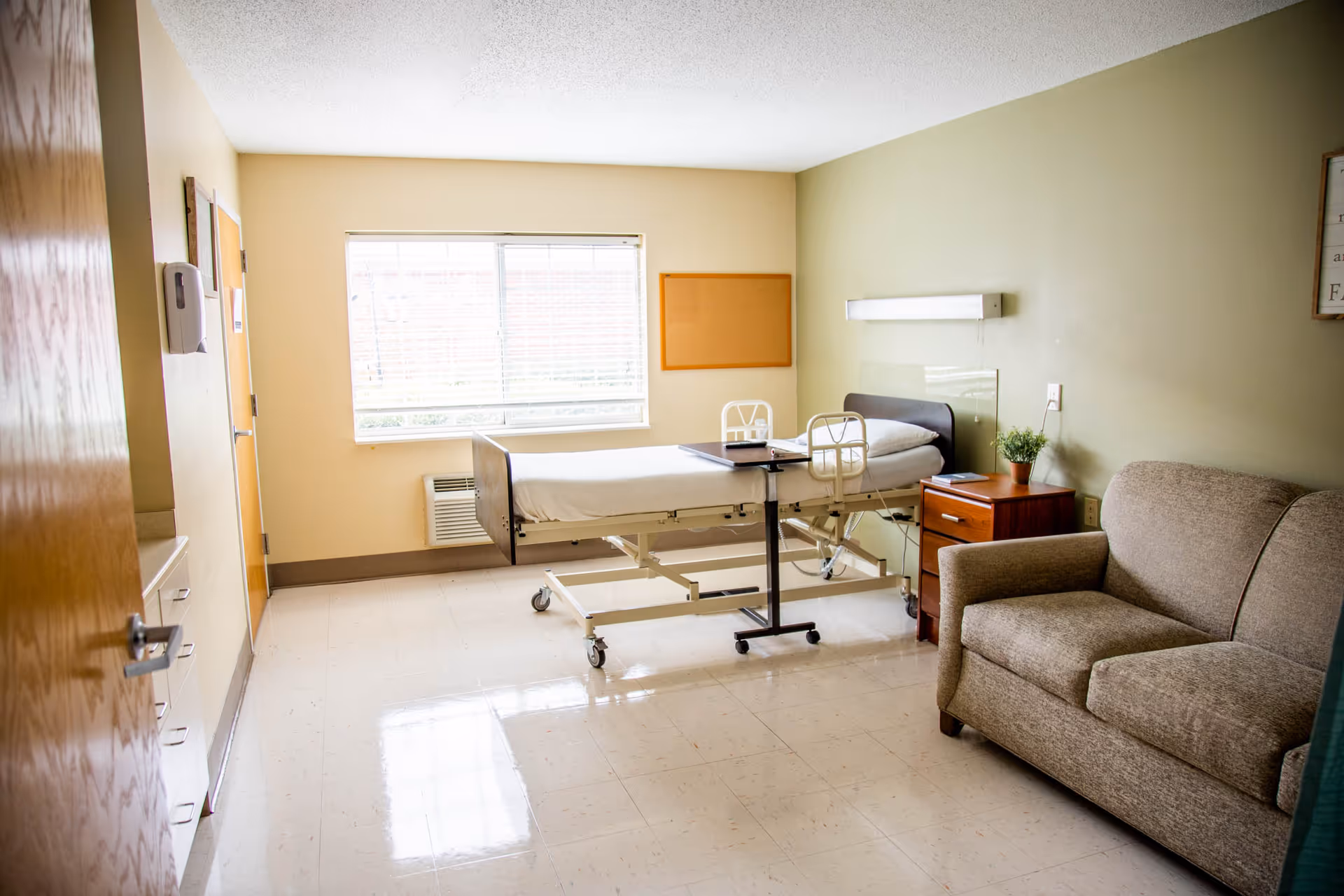 A bright and clean nursing facility room with a hospital bed, a small wooden nightstand with a plant and book, a beige upholstered loveseat, and a large window with blinds letting in natural light. The walls are painted in soft yellow and green tones.