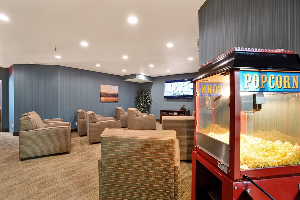A senior living lounge with upholstered armchairs arranged facing a wall-mounted TV and a popcorn machine in the foreground.