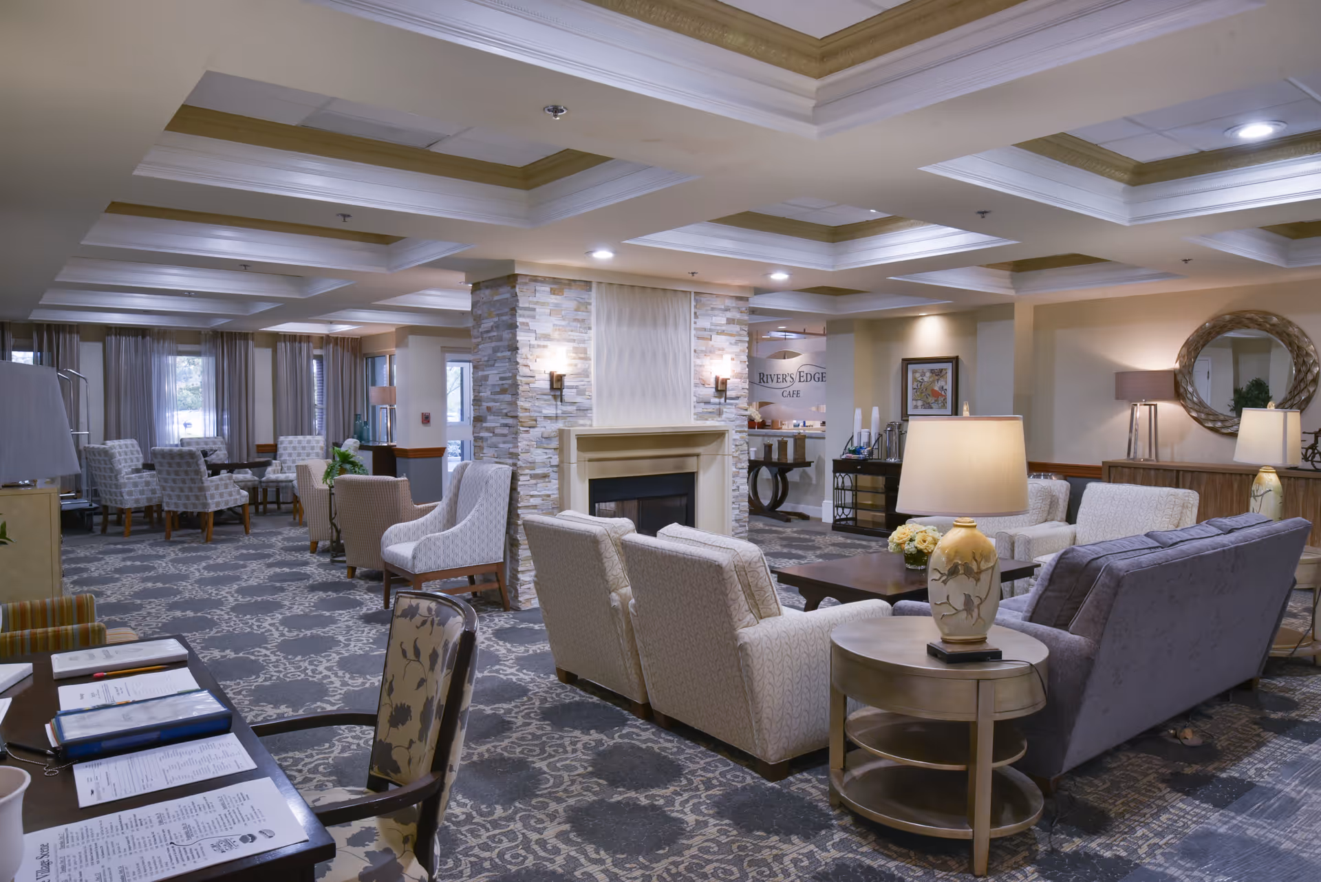 A spacious and well-lit senior living facility common area with comfortable seating including armchairs and sofas arranged around a central fireplace. The room features patterned carpet, multiple table lamps, a round mirror on the wall, and a sign indicating the Rivers Edge Cafe in the background.