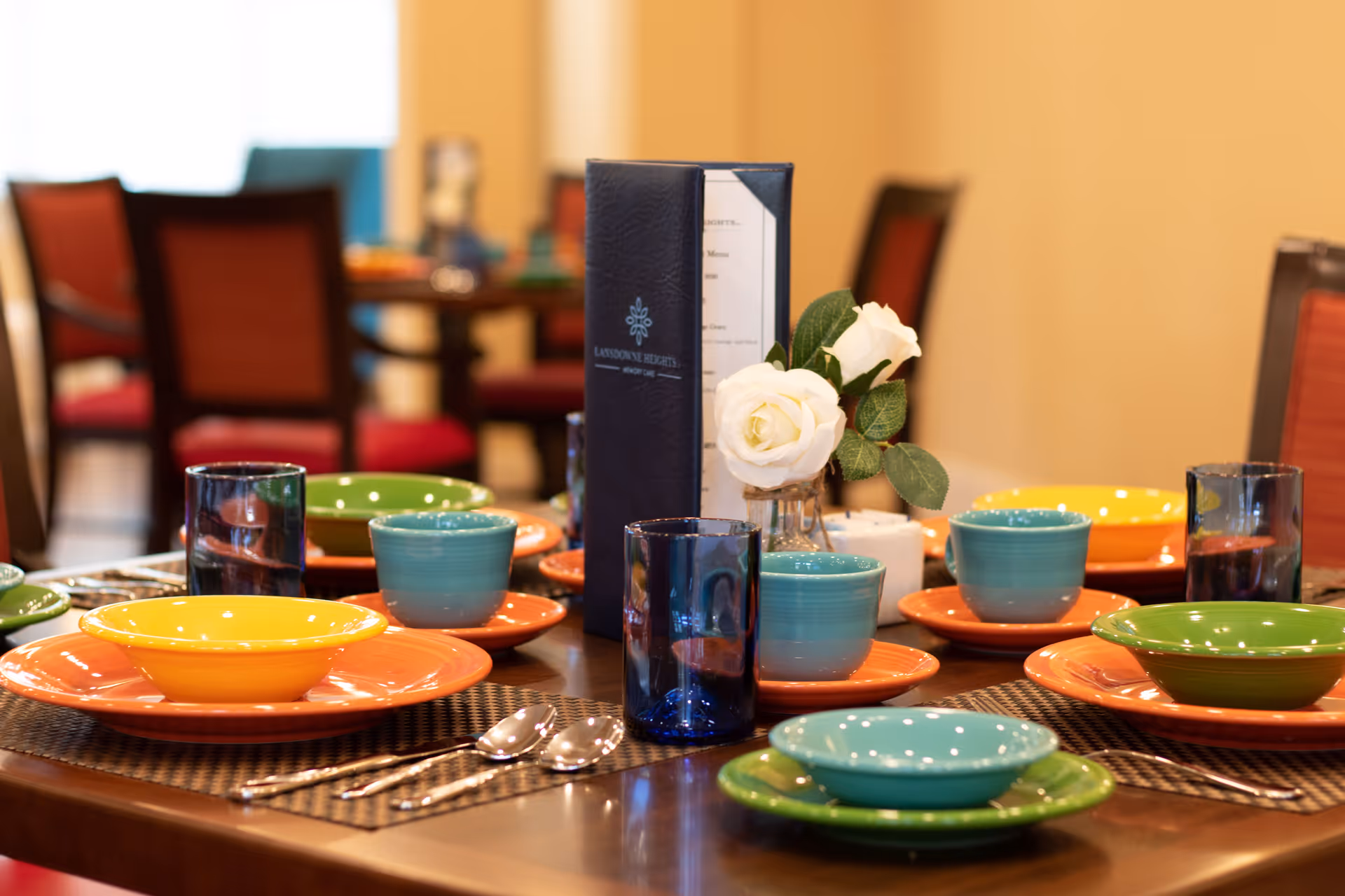 A dining table set with colorful plates, bowls and blue glasses, a menu holder and a small vase of white roses.
