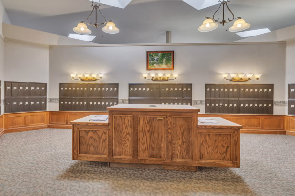 Interior view of a mailroom with multiple rows of mailboxes mounted on the walls. There is a large wooden central table with papers on it. The room has carpeted flooring, soft wall lighting, and two ceiling light fixtures. A framed painting hangs on the wall above the mailboxes.
