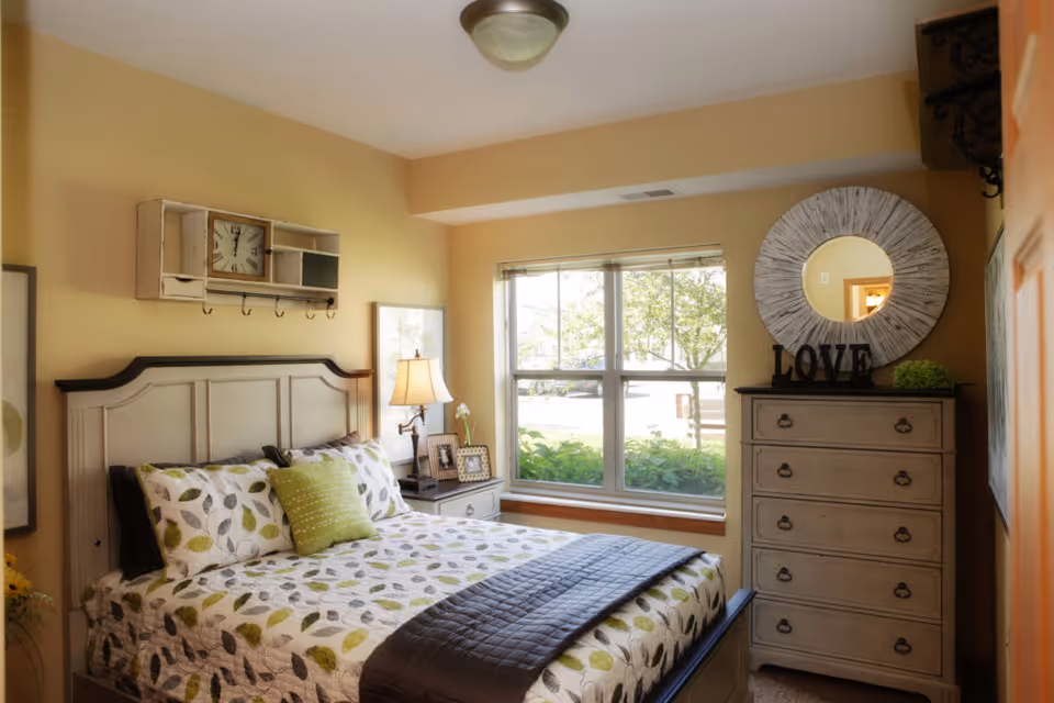 A cozy bedroom with a bed featuring a leaf-patterned quilt and green accent pillow. There is a nightstand with a lamp and framed photos next to the bed. A large window lets in natural light, and a dresser with a round decorative mirror and the word 'LOVE' on top is against the wall.