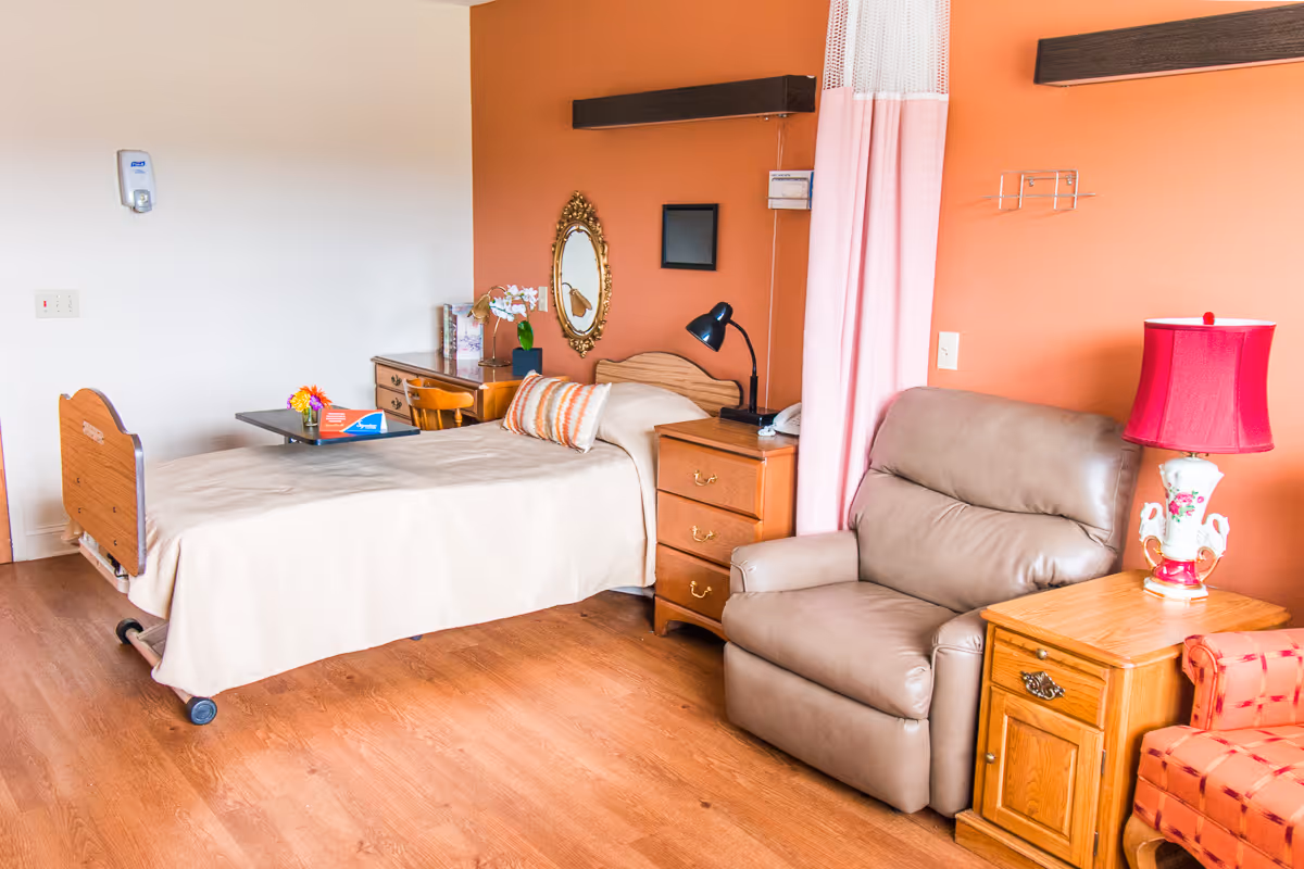 A senior living facility bedroom with a single bed covered in a beige blanket, a wooden nightstand with a black lamp, a beige recliner chair, a wooden side table with a decorative lamp with a red shade, and a small desk with a chair. The walls are painted white and orange, and there is a decorative mirror and a small picture frame on the orange wall.