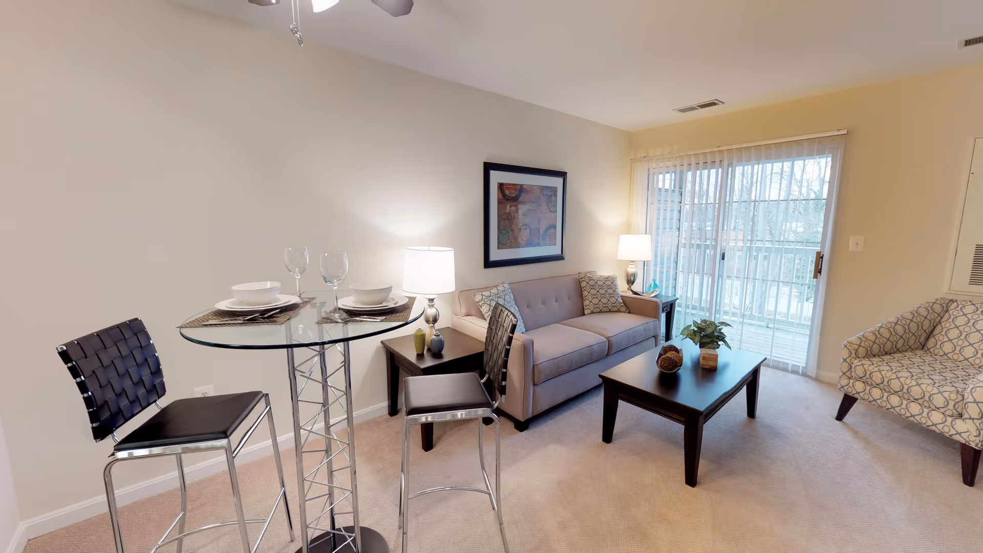A cozy living room area with a beige sofa, a patterned armchair, a dark wooden coffee table with decorative items, and a glass-top dining table with two black woven chairs. The room has beige carpet, a ceiling fan, a framed artwork on the wall, a side table with a lamp, and sliding glass doors with vertical blinds leading to a balcony.