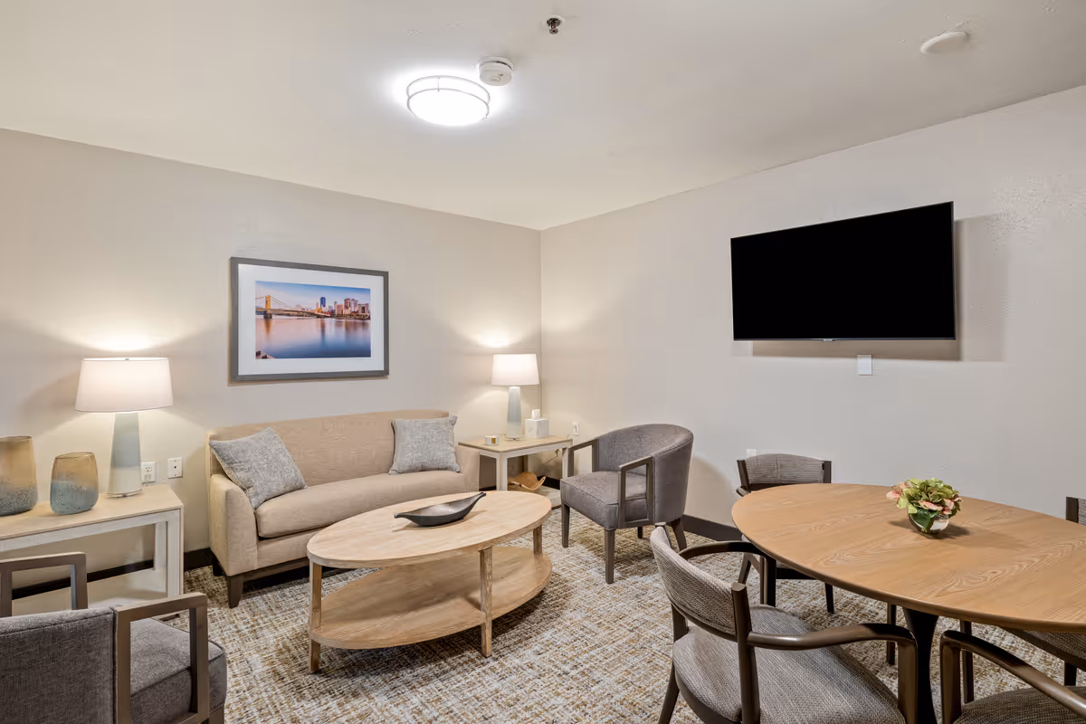 A cozy living room with a beige sofa, two gray armchairs, a wooden oval coffee table, and a round wooden dining table with four chairs. The room is softly lit by two table lamps on side tables and a ceiling light. A flat-screen TV is mounted on the wall, and a framed picture of a cityscape hangs above the sofa.