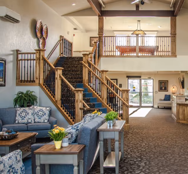 A spacious and well-lit common area in an assisted living facility featuring a staircase with wooden railings and carpeted steps. The room includes comfortable blue sofas with patterned cushions, wooden side tables with potted plants and flowers, and a large windowed entrance door letting in natural light. The upper level has a railing and a hanging light fixture, and the overall decor is warm and inviting.
