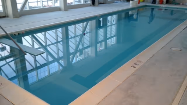 Indoor swimming pool with clear blue water, surrounded by a tiled deck and large windows reflecting on the water surface.