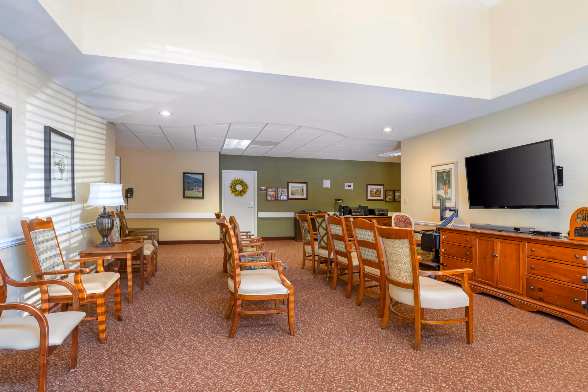 A spacious common area with multiple wooden chairs arranged in rows facing a large flat-screen TV mounted on the wall. The room has beige walls, carpeted floor, framed artwork, a wooden sideboard under the TV, and a table lamp on a small table. There is a door with a decorative wreath and a green accent wall in the background.