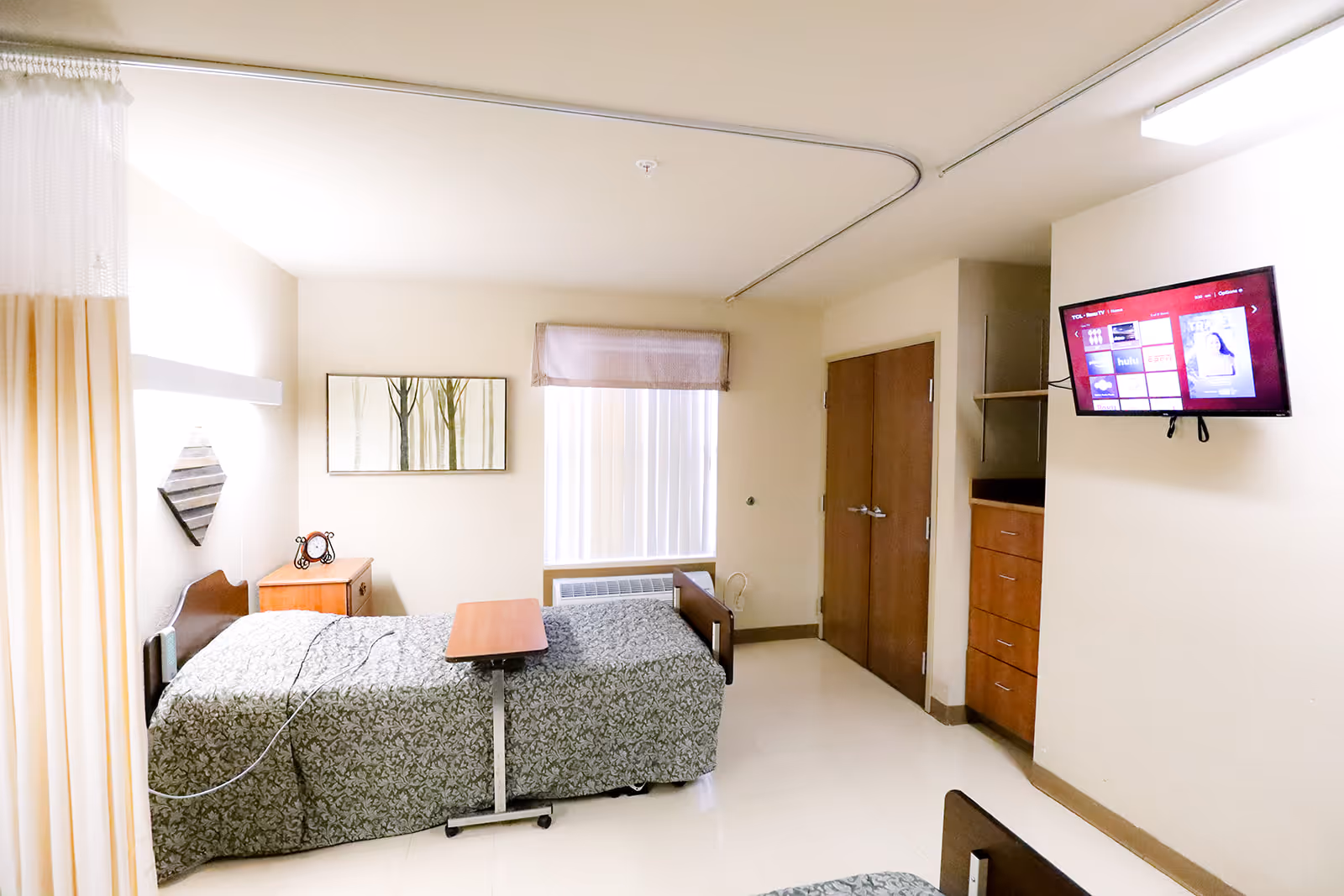 A simple, clean bedroom in a healthcare facility with a single bed covered in a patterned bedspread, a small wooden bedside table with a clock, a wall-mounted TV, a window with vertical blinds and a valance, and a wooden closet with double doors. The room has light-colored walls and tiled flooring.